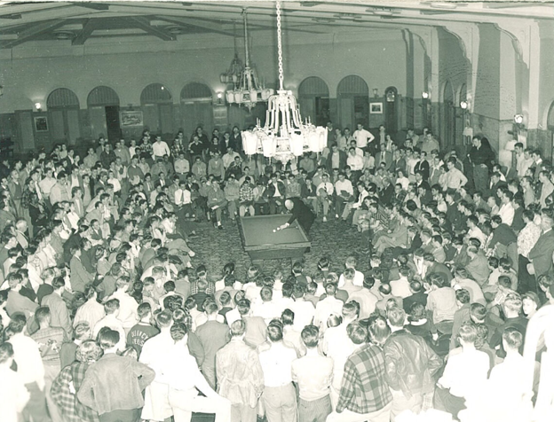 Prompt A. A large group of students gather inside a ballroom to watch a man playing pool.