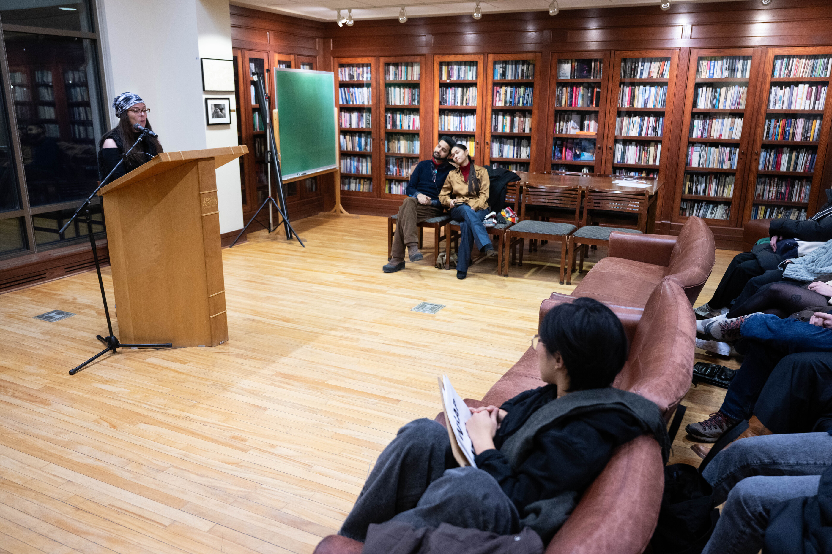 Prompt B. A person reads from a podium during an Iowa Writers' Workshop event as audience members listen in a library.